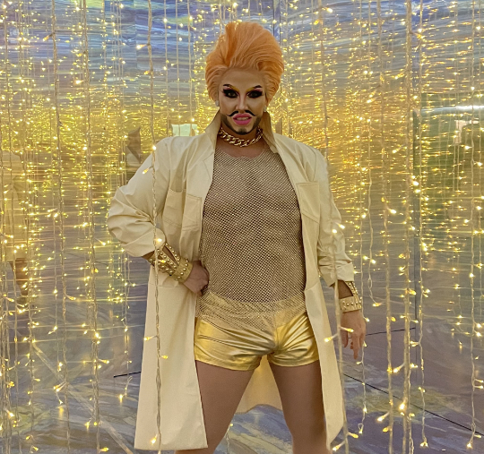 A Drag King dressed fabulously in golden glitter stands in a shiny room